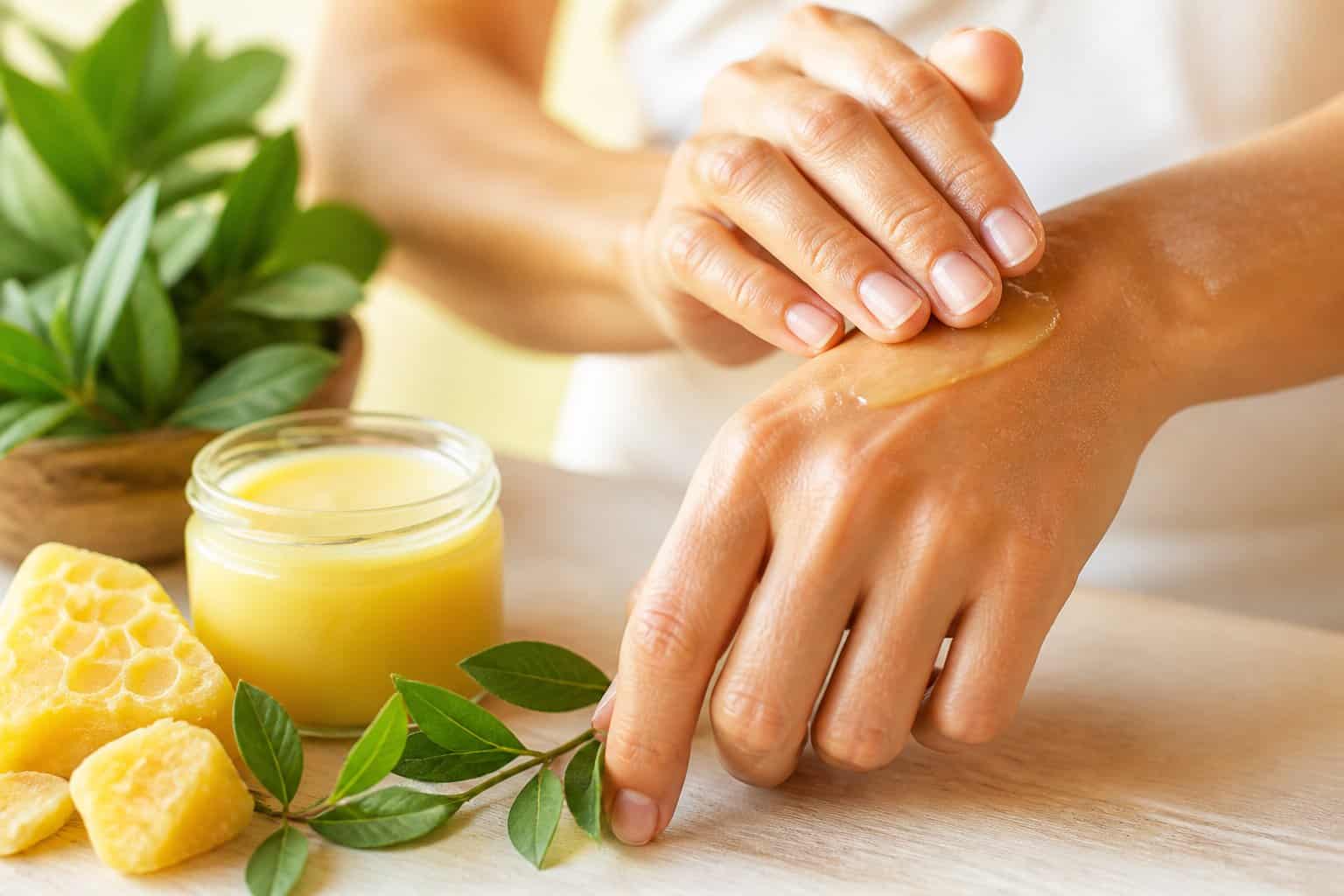 Close-up of hands applying beeswax balm onto skin with a jar of beeswax cream and natural beeswax pieces nearby.