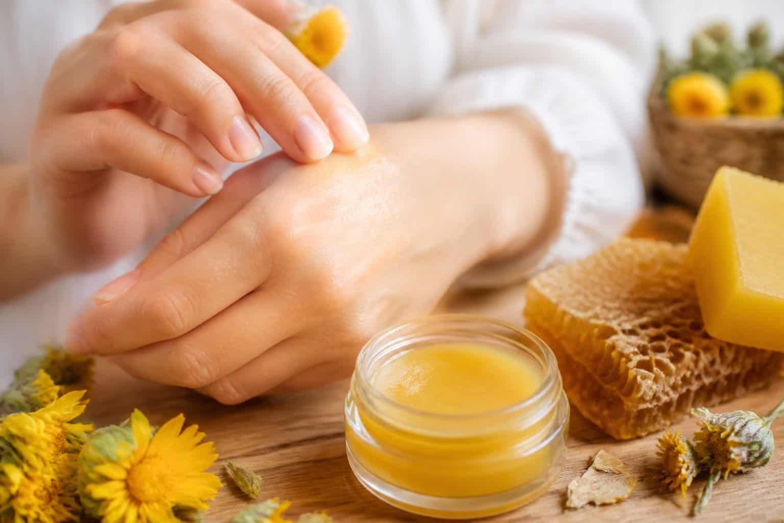 Close-up of hands applying beeswax balm to skin with a jar of beeswax and honeycombs nearby.
