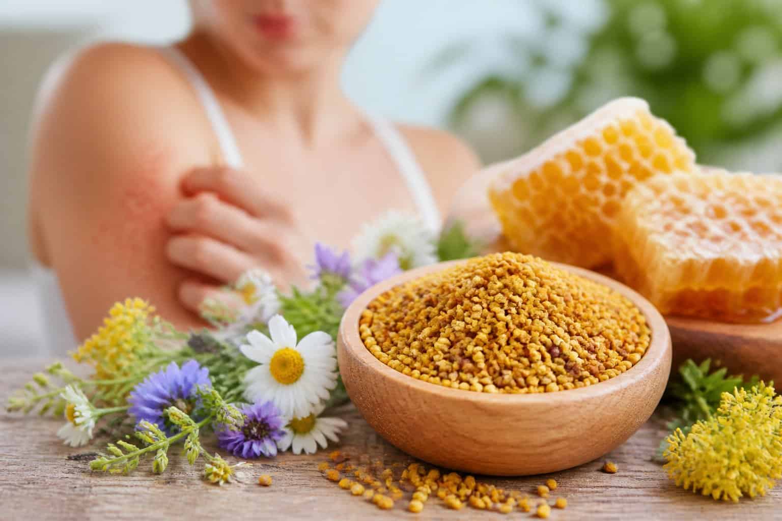 A bowl of bee pollen granules with wildflowers and honeycomb nearby, and a person softly touching their skin in the background.